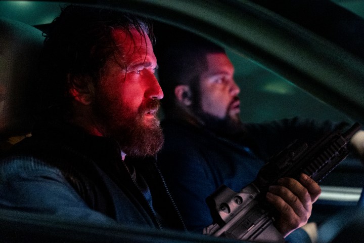 Gerard Butler holds a gun while sitting in the front seat of a car.