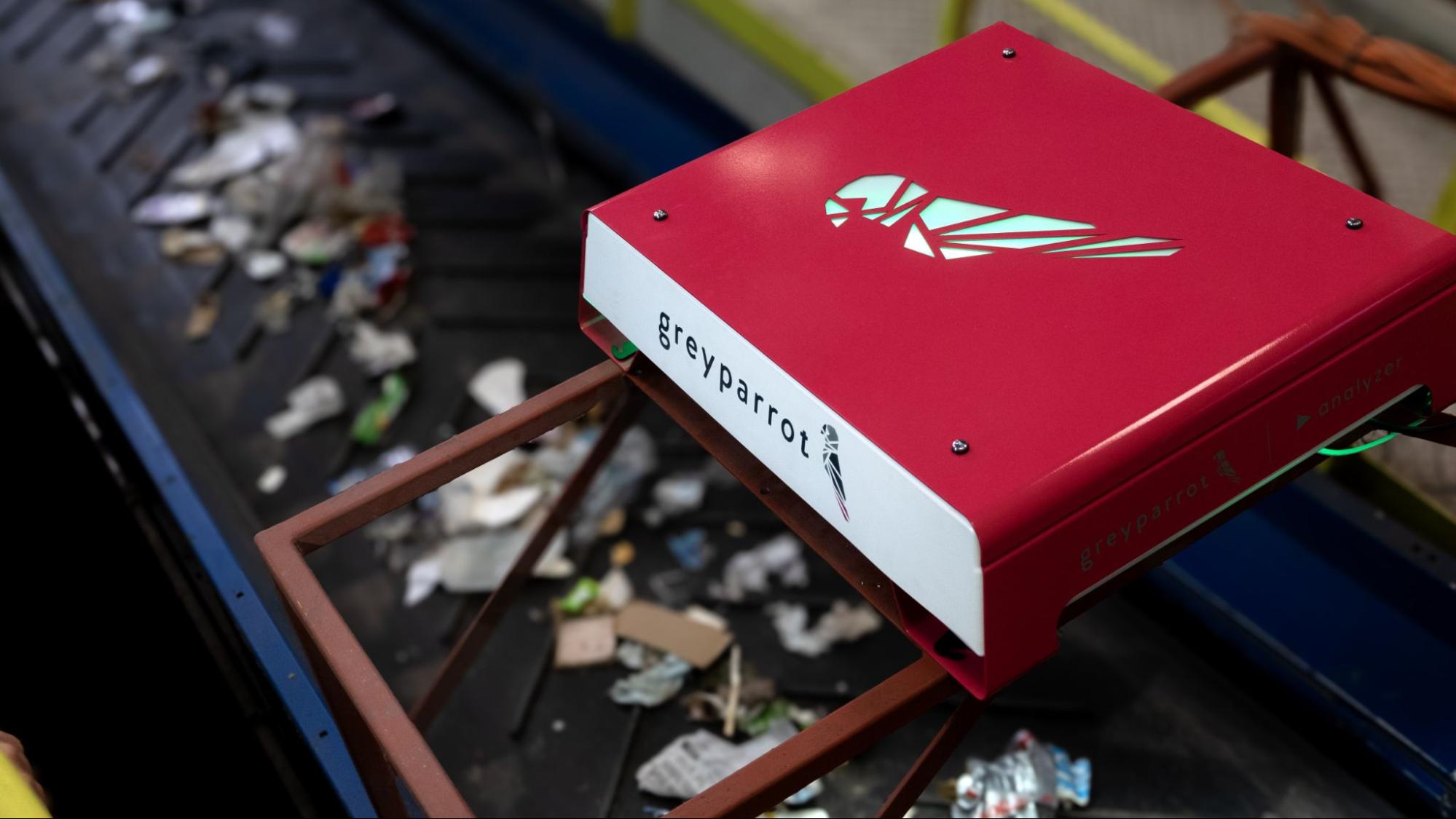 A bright red and white metal box is attached to a metal frame and sits above a conveyor belt carrying waste products.