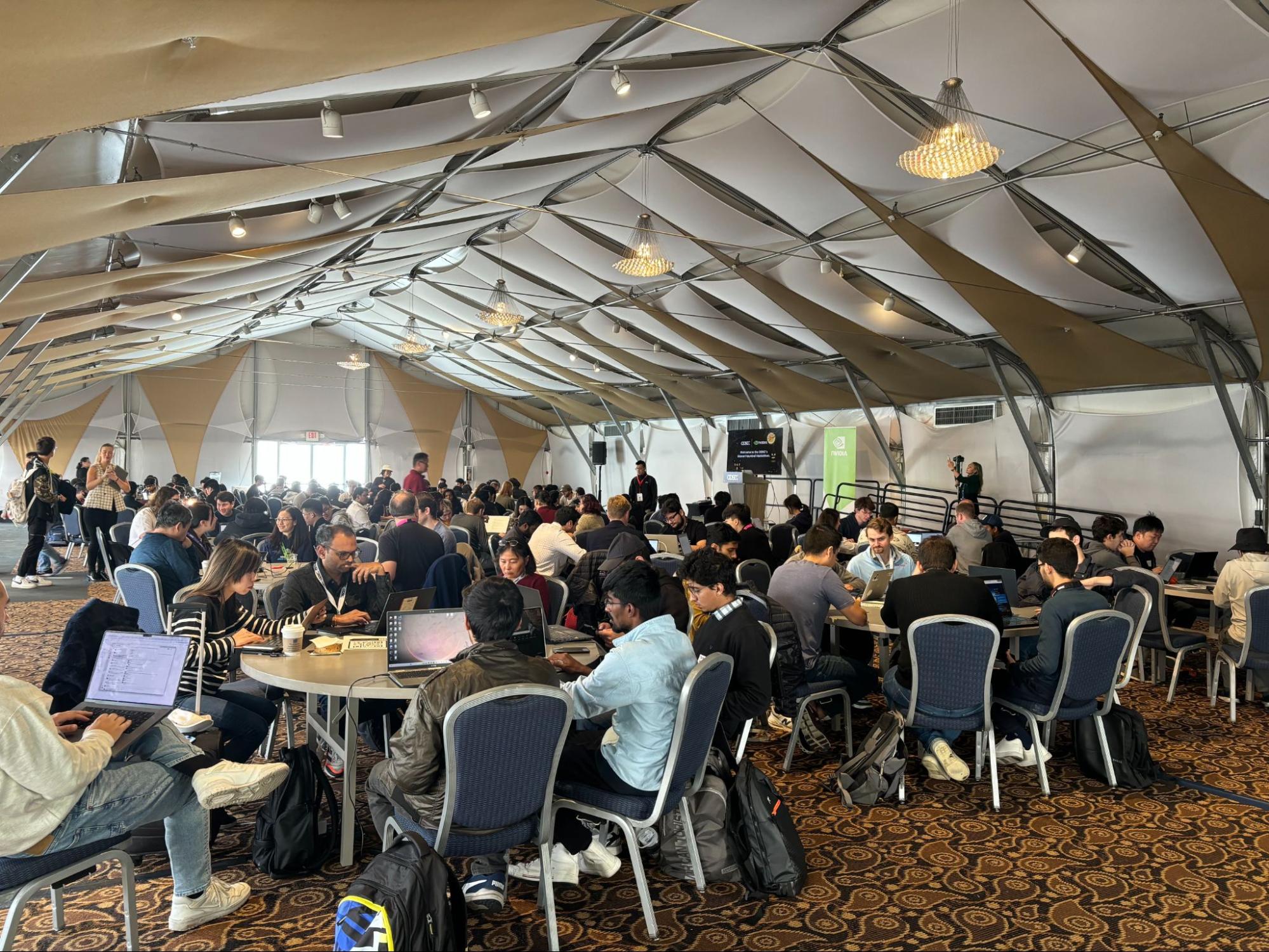 Photo of NVIDIA hackathon participants sitting around tables working on laptops.
